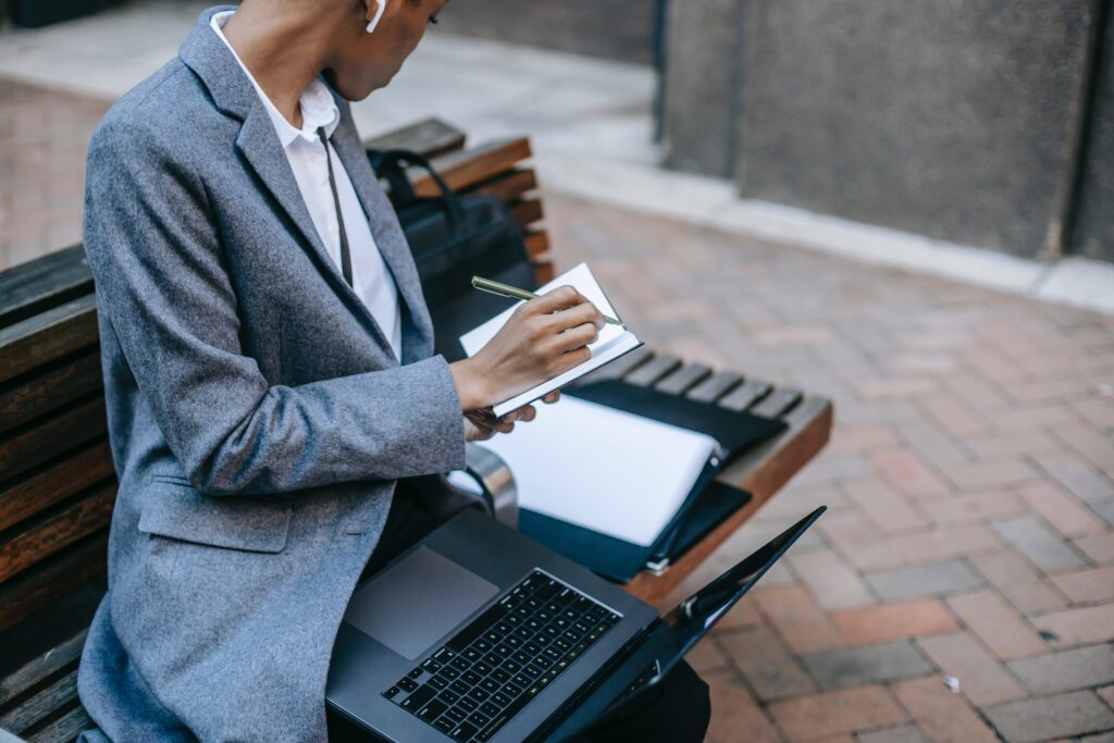 woman financial professional in suit, sitting on park bench with laptop while taking notes