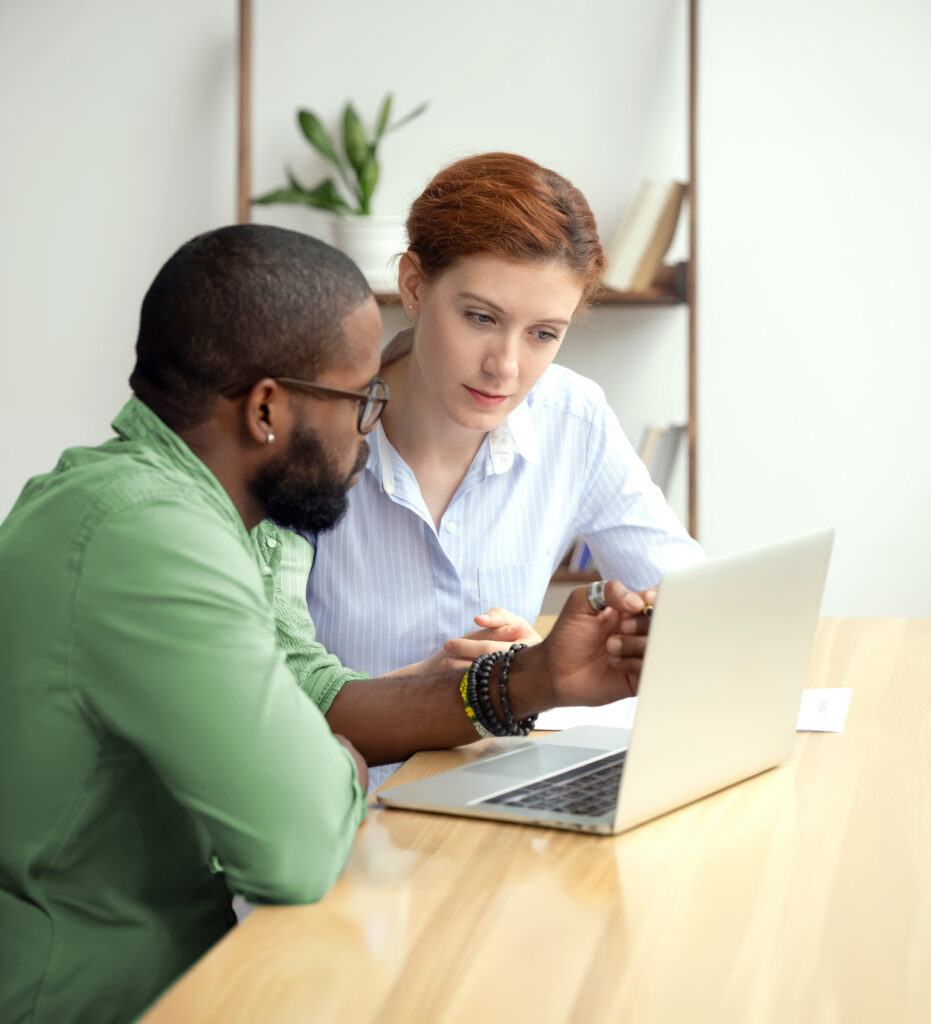Young man with glasses sitting at a laptop and pointing at the screen while talking with a female coworker in office setting