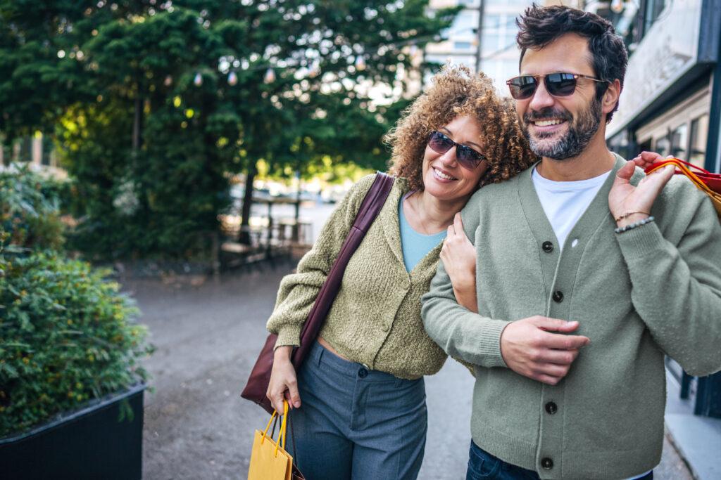 Young couple walking hand in hand down a bustling street, carrying colorful shopping bags and radiating joy after enjoying a delightful shopping spree together